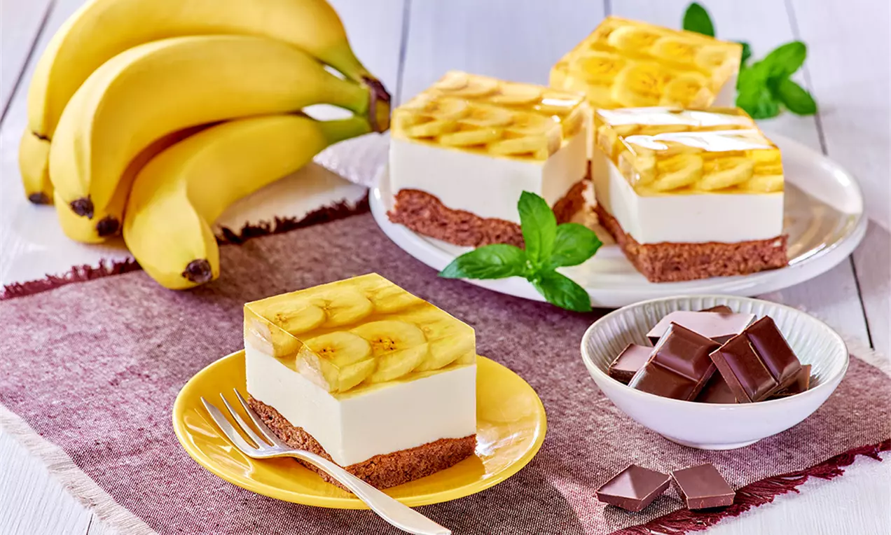 Kvadrato formos desertas su kakaviniu pagrindu, baltu kremu, želė viršuje, papuoštas bananų griežinėliais, pateiktas ant geltonos lėkštės su šakute.