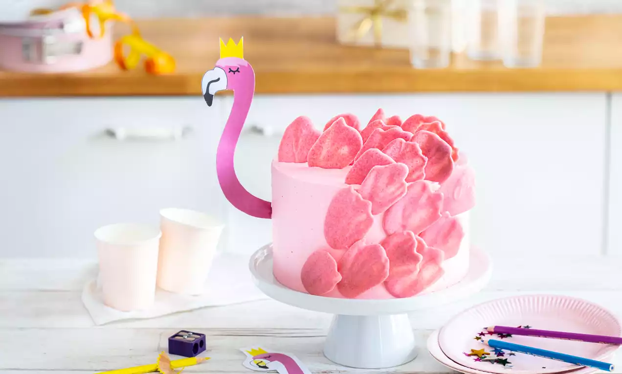 Šviesiai rožinis apvalios formos tortas su cukraus lapeliais, sudėliotais kaip flamingo plunksnos, dekoruotas popierine flamingo galva ant balto stovo