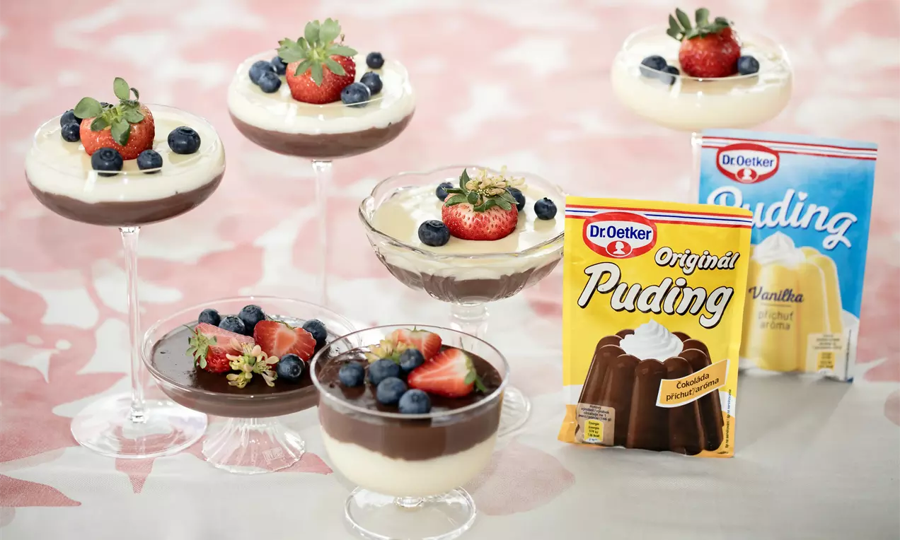 Dviejų sluoksnių pudingas taurėse: šokolado ir vanilės, viršuje mėlynės, braškės ir eglės šakelė, dekoruotas ant pastelinių atspalvių staltiesės