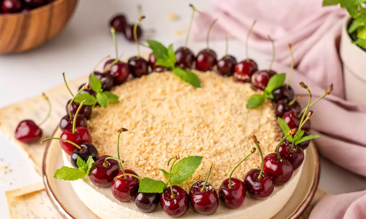Šviesiai kreminis tortas su švytinčios geltonos trupiniais viršuje, apsuptas sodrių vyšnių vainiku ir žalumynais ant paprastos baltos lėkštės