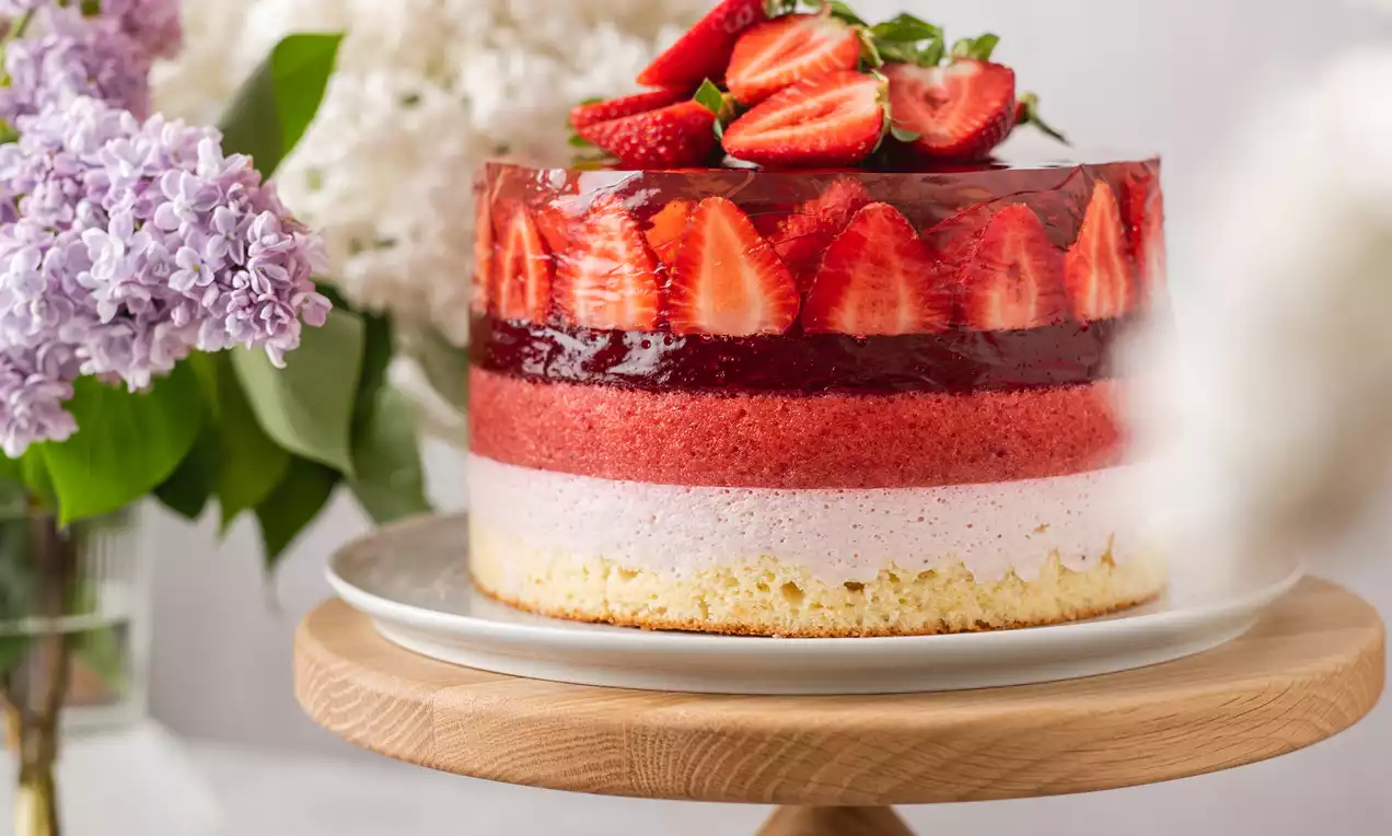 Ryškiai rožinis tortas su sluoksniais kreminės ir uoginės tekstūros, puoštas šviežiomis braškėmis ant medinio padėklo